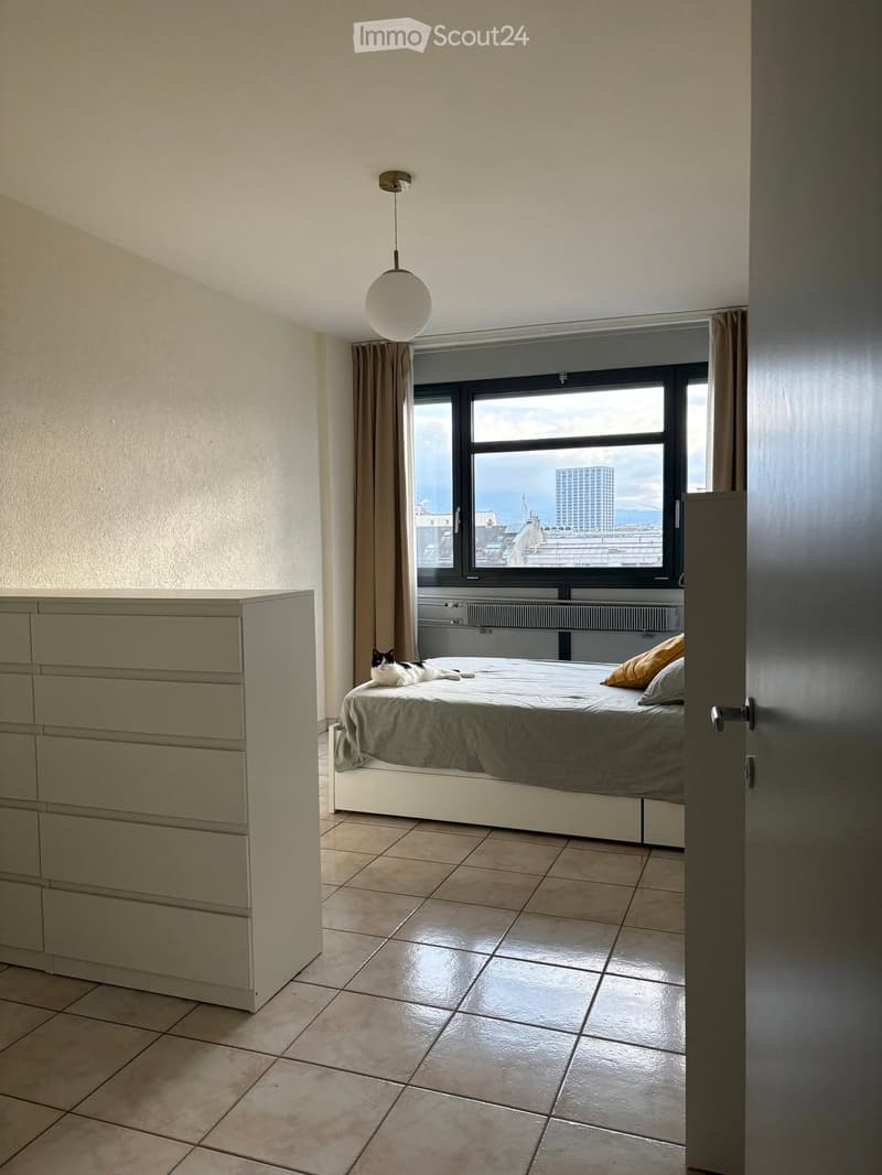 Modern bedroom with tiled floor, large window offering city view, bed with pillows, dresser, and a cat resting on the bed.