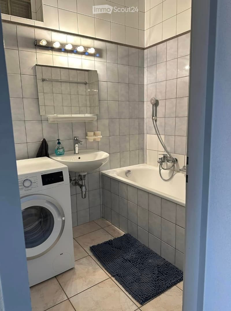 Modern bathroom with a white sink, bathtub, washing machine, and a dark blue bath mat on the floor.