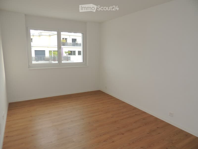 Empty room with wooden flooring and white walls, featuring a large window with a view of a neighboring building and balcony.