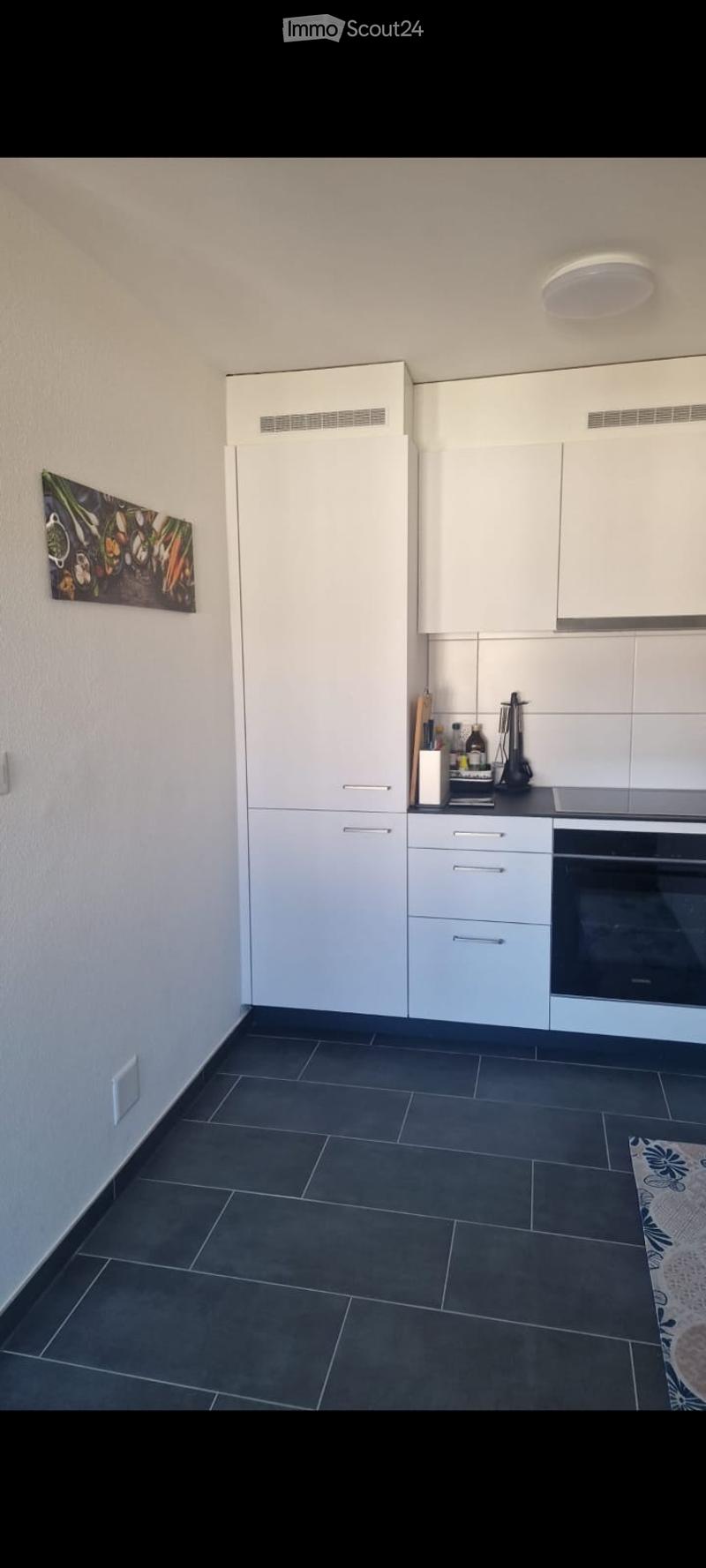 Modern kitchen with white cabinets, dark tiled floor, and a black oven. A small cutting board and a few bottles are placed on the counter, and a picture hangs on the wall.
