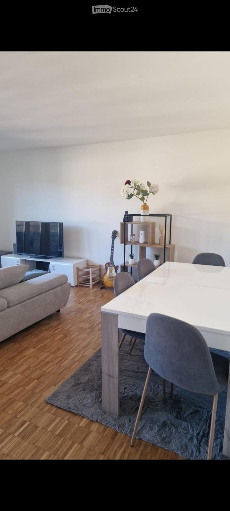 Modern living and dining area with a grey sofa, white dining table, grey chairs, a guitar, a shelf with decorative items, and a television.