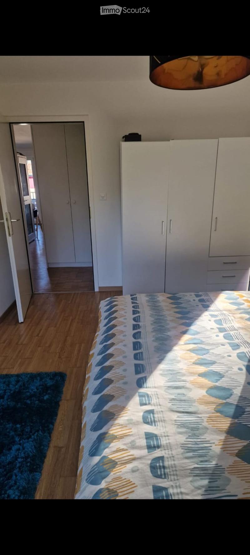 Bright bedroom with wooden flooring, a bed with a blue and yellow patterned duvet, a white wardrobe, and a blue rug.