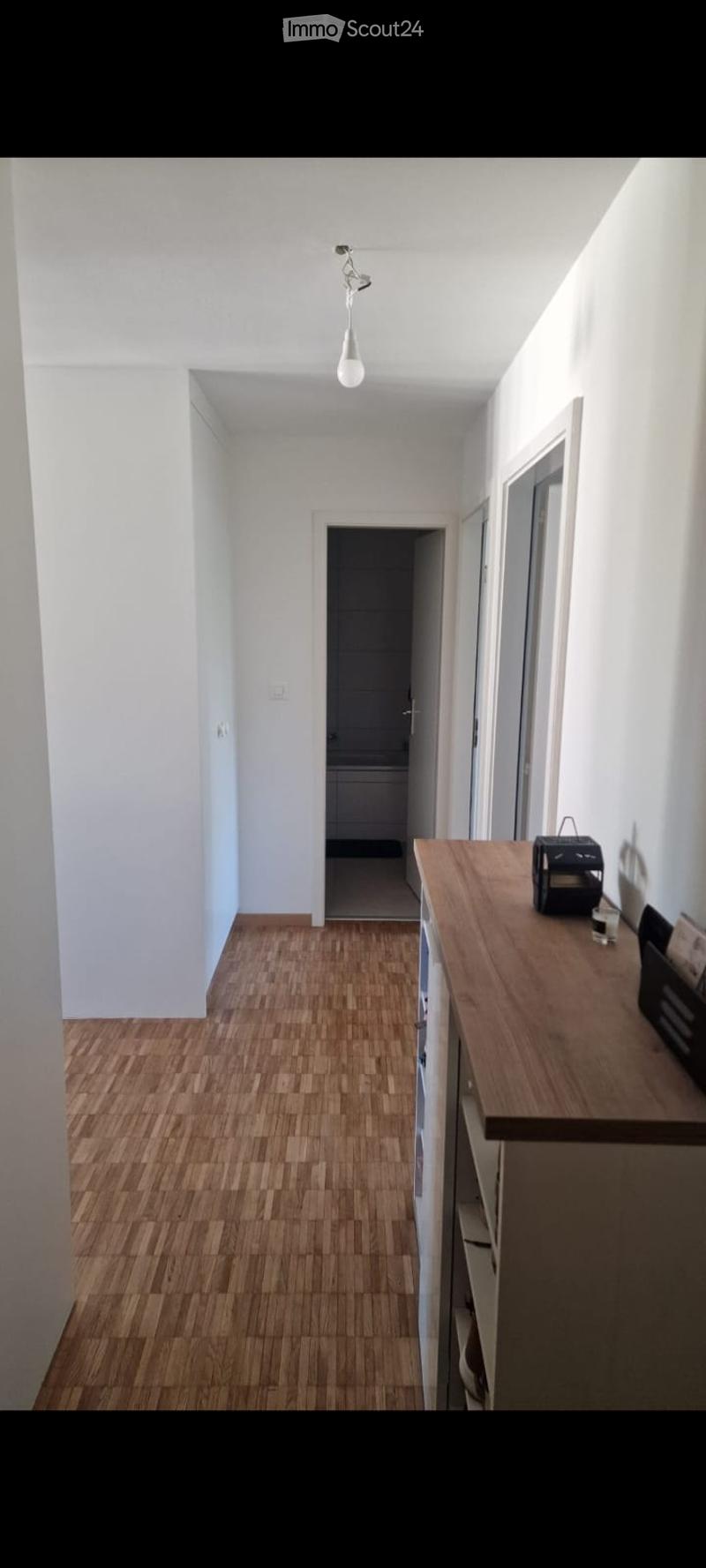 A narrow hallway with wooden flooring, white walls, and a single light fixture. A small table with a few items sits on the right side, and a doorway leads to another room.