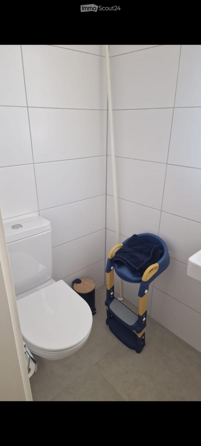 Modern bathroom with white tiled walls, a toilet, a small wooden stool, and a blue step ladder with a towel on top.