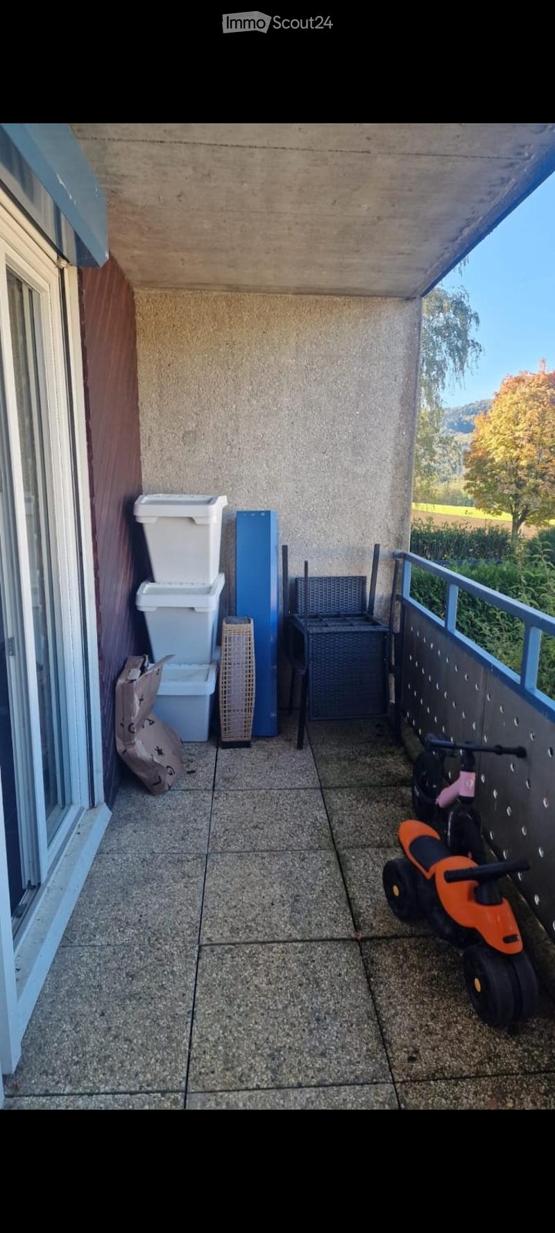 Balcony with tiled floor, metal railing, and a view of greenery. Contains storage bins, a wicker chair, and a small toy car.
