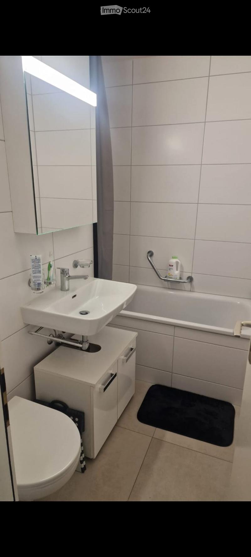 Modern bathroom with white tiles, a sink with a modern faucet, a mirror, a toilet, a bathtub with a handrail, and a small cabinet under the sink.