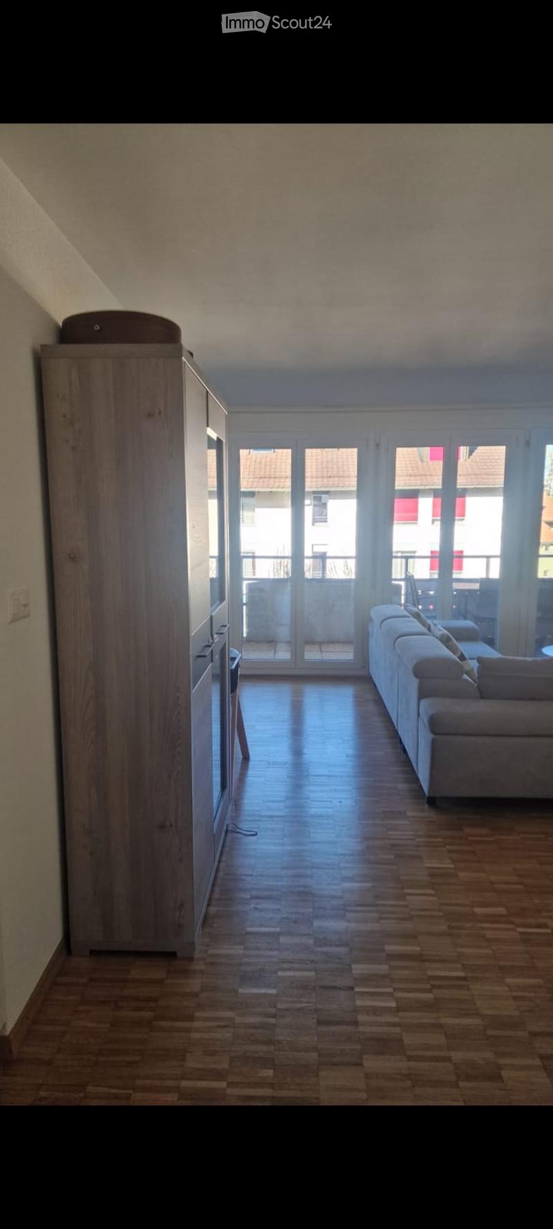 Bright living room with wooden flooring, large windows, and a balcony access. Features a grey sofa and a tall wooden cabinet.