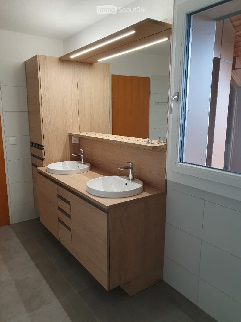 Modern bathroom with two white sinks, wooden cabinetry, large mirror with integrated lighting, and sleek chrome faucets.