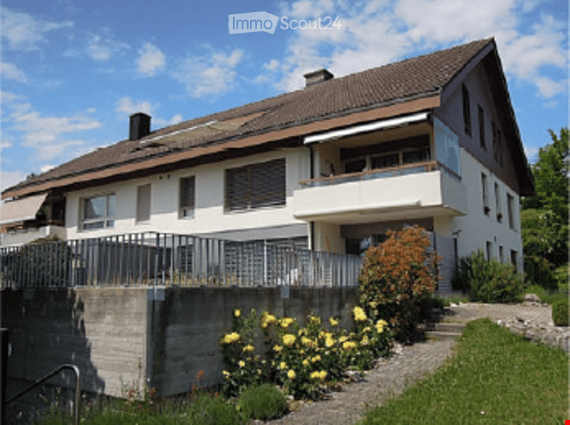 Two-story house with a brown tiled roof, white walls, multiple windows, and a balcony on the upper floor. The house is surrounded by a well-maintained garden with colorful flowers and shrubs.