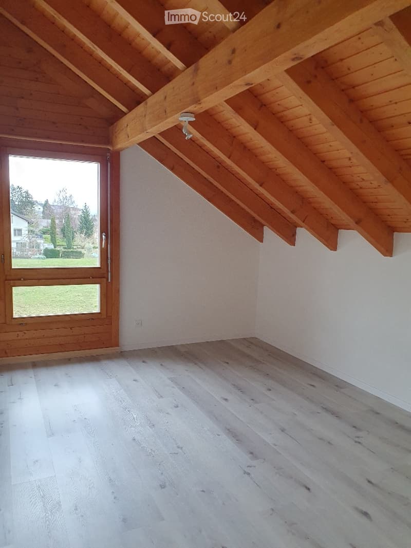 Empty attic room with wooden beams, white walls, and light wooden flooring. Features a large window with a view of the outdoors.