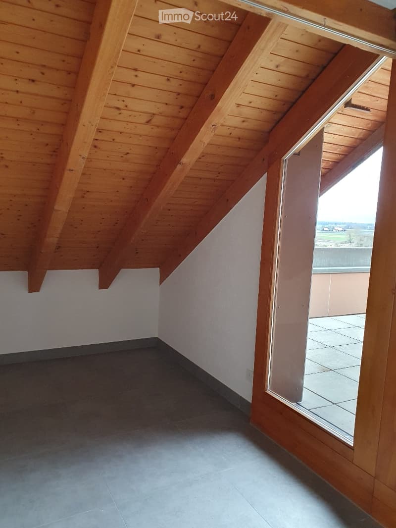 Empty attic space with wooden beams, exposed wooden ceiling, grey tiled floor, and large window opening to a balcony.