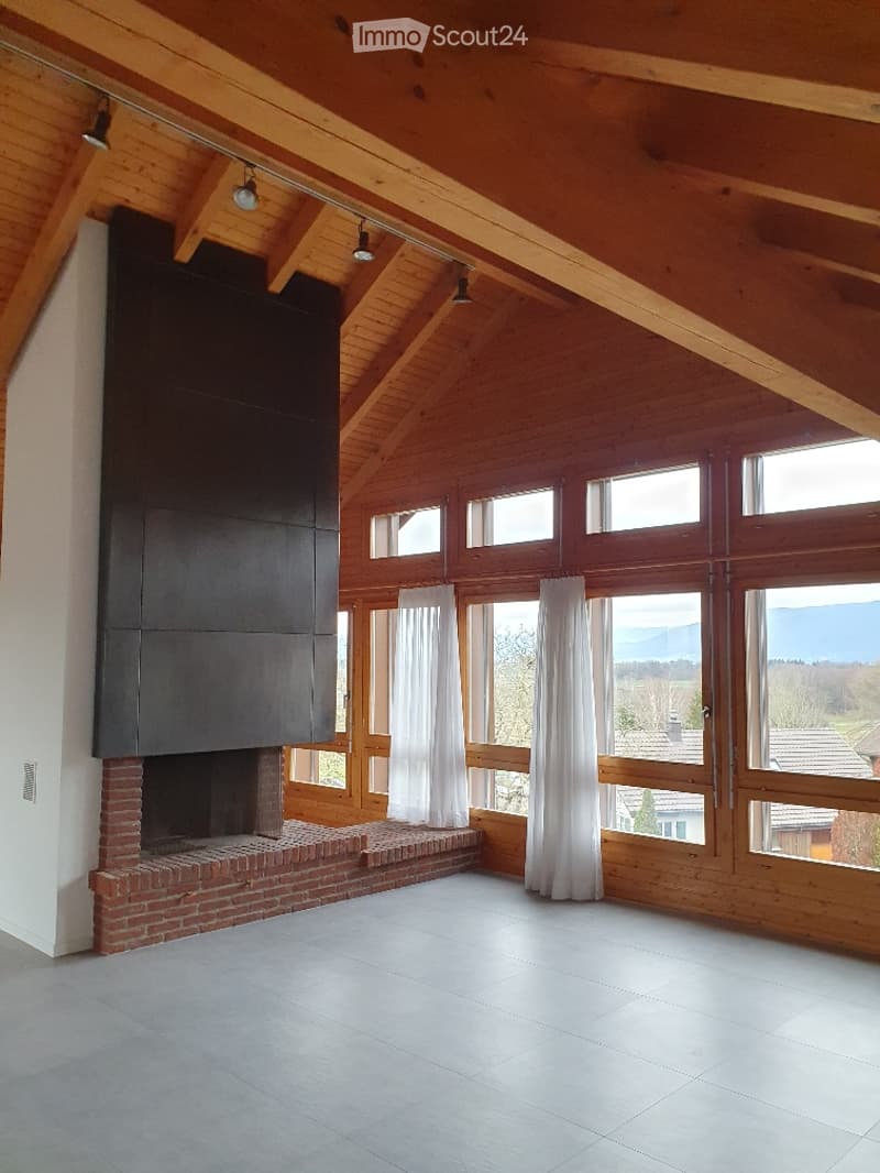 Bright, open room with exposed wooden beams, large windows offering a scenic view, and a brick fireplace with a modern black surround.