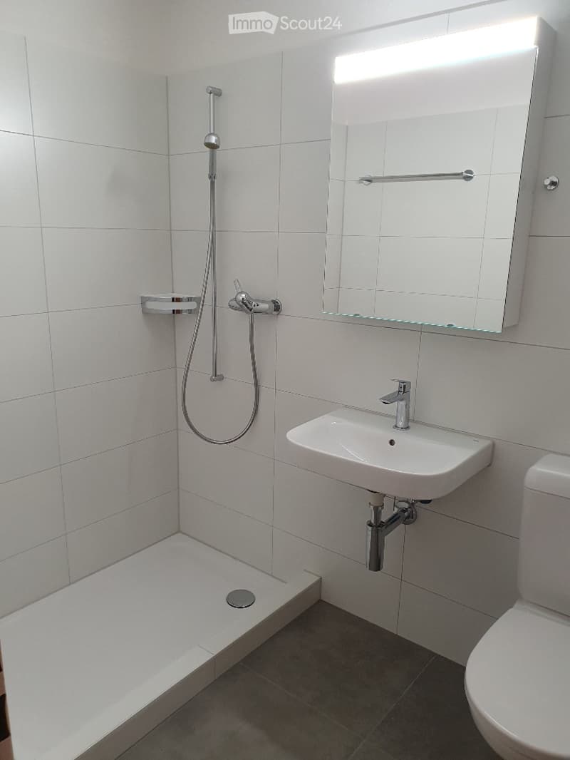 Modern bathroom with white tiled walls, a corner shower, a white sink with a chrome faucet, a toilet, and a large illuminated mirror.