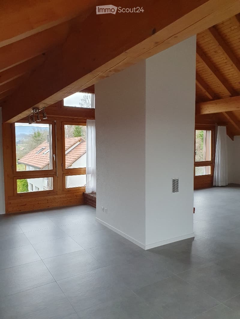 Bright, open attic space with wooden beams, large windows offering a scenic view, and a smooth tiled floor.
