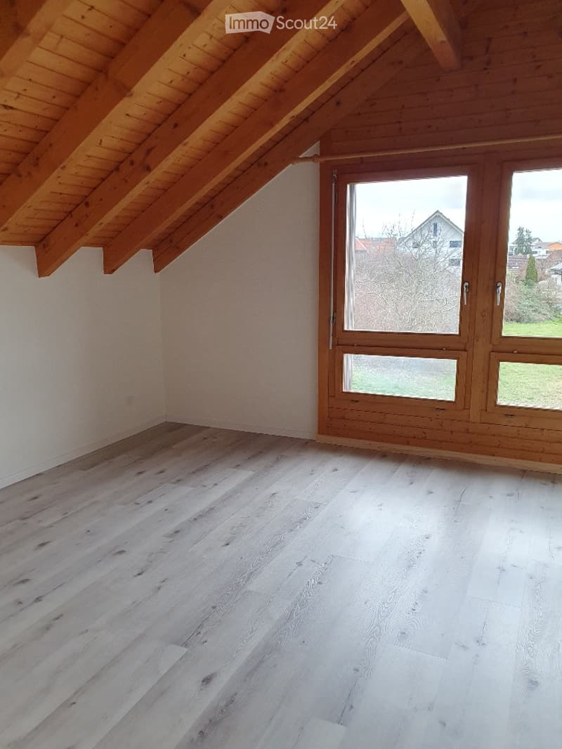 Empty attic room with wooden slanted ceiling, light wooden flooring, and large windows with wooden frames.