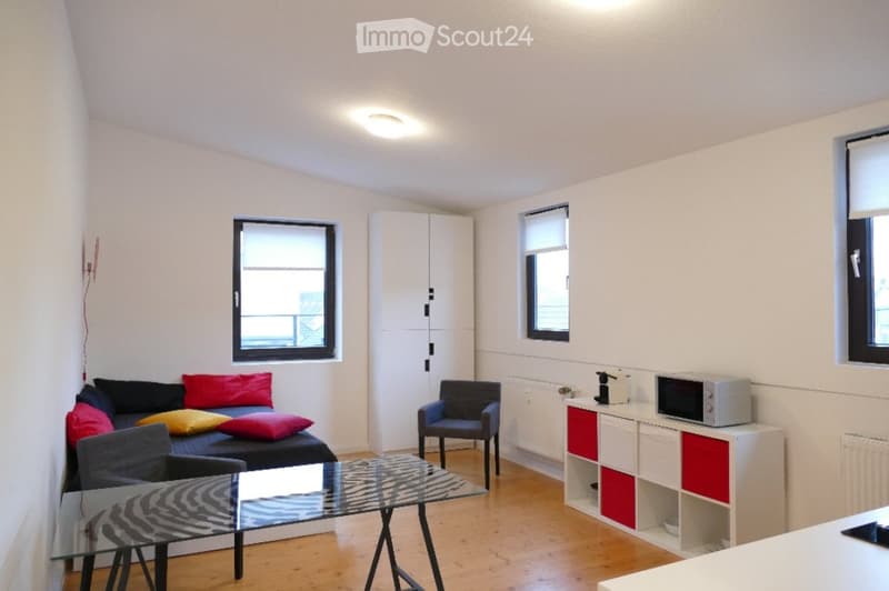 A modern, compact living space with a sofa, colorful pillows, a glass coffee table, a chair, a white cabinet, and a microwave on a small counter.