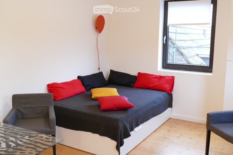 A small, modern living room with a grey sofa, colorful pillows, a glass coffee table, and a view of the roof from a large window.