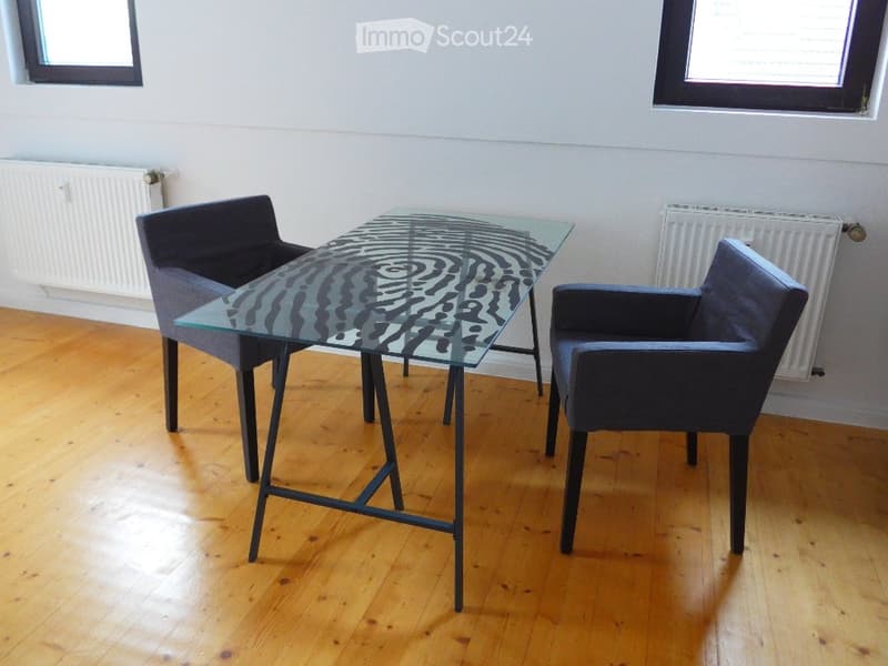 A modern, minimalist room with a glass table and two dark blue chairs, wooden flooring, and white walls.