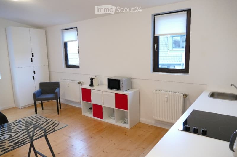 A modern, compact kitchen and living area with white walls, wooden flooring, and large windows. Features include a white cabinet with red accents, a microwave, a sink, an induction stove, a glass table, and a blue chair.