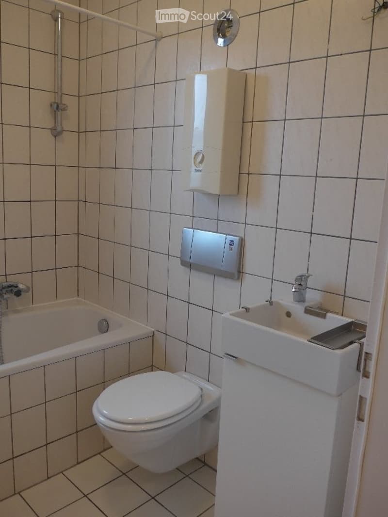Modern bathroom with white tiled walls and floor, featuring a toilet, bathtub, sink with a chrome faucet, and a showerhead with a rail.