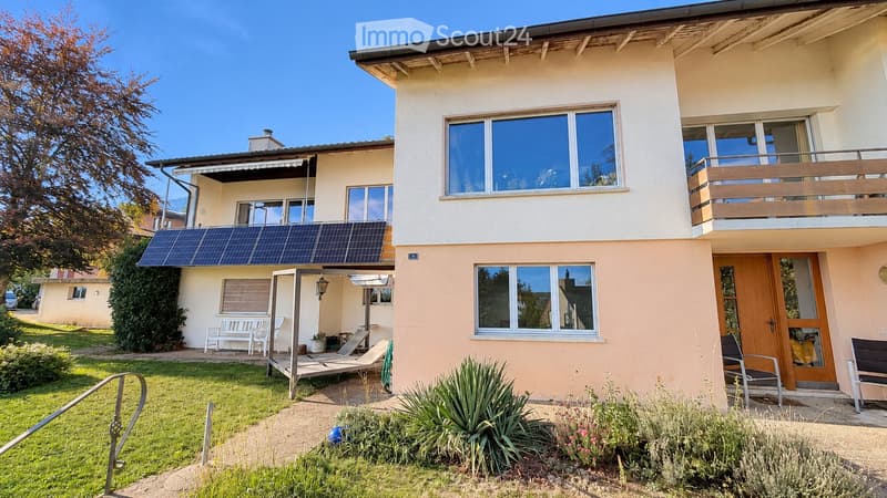 Two-story house with solar panels on the roof, large windows, a balcony, and a well-maintained garden with outdoor seating.