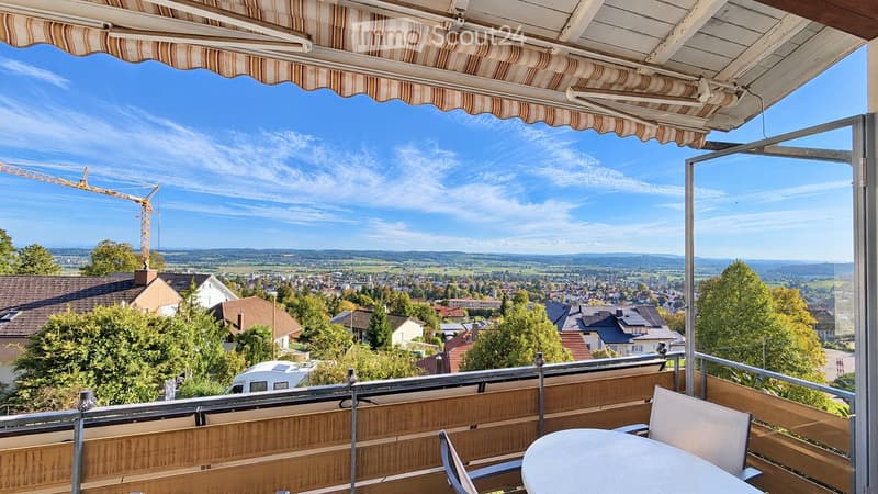 Balcony with a view of a town and countryside, featuring a table and chairs under a striped awning.