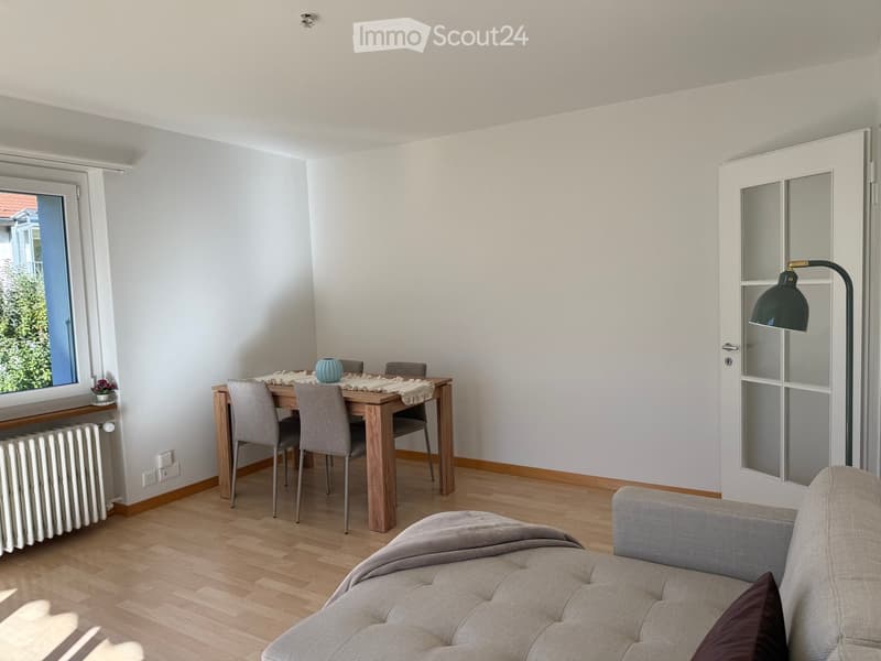 A modern, well-lit living and dining area with light wooden flooring, white walls, and large windows. Features a grey sofa, a wooden dining table with four chairs, a decorative vase, and a floor lamp near the door.