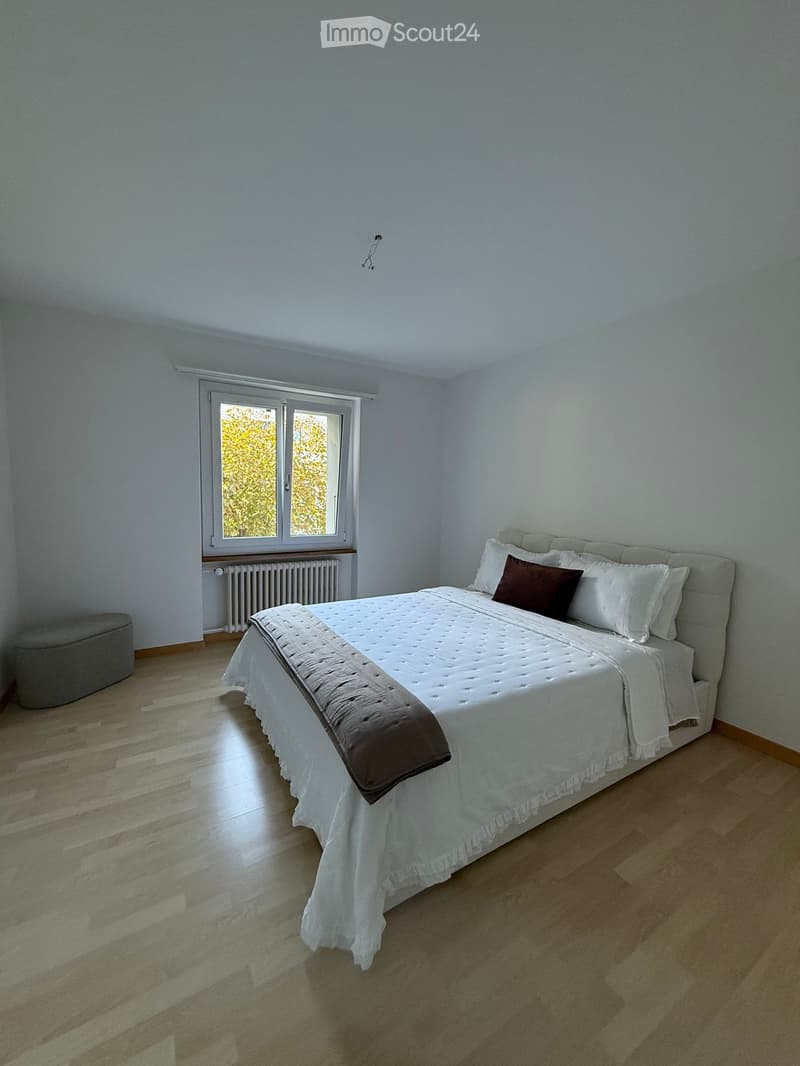 A well-lit bedroom with a large bed, white bedding, and a grey stool near the window. The room features light wooden flooring and white walls.
