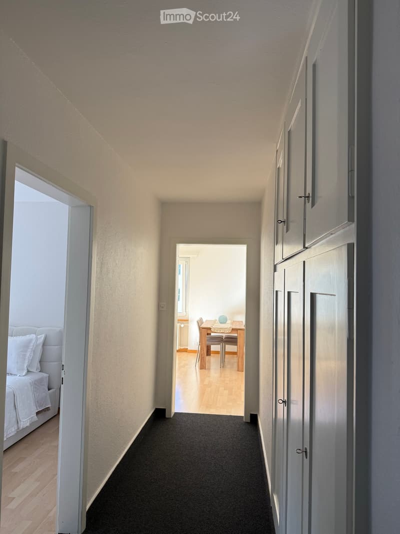 A narrow hallway with light-colored walls, carpeted floor, and white doors leading to adjacent rooms. Visible rooms include a bedroom with a white bed and a dining area with a wooden table and chairs.