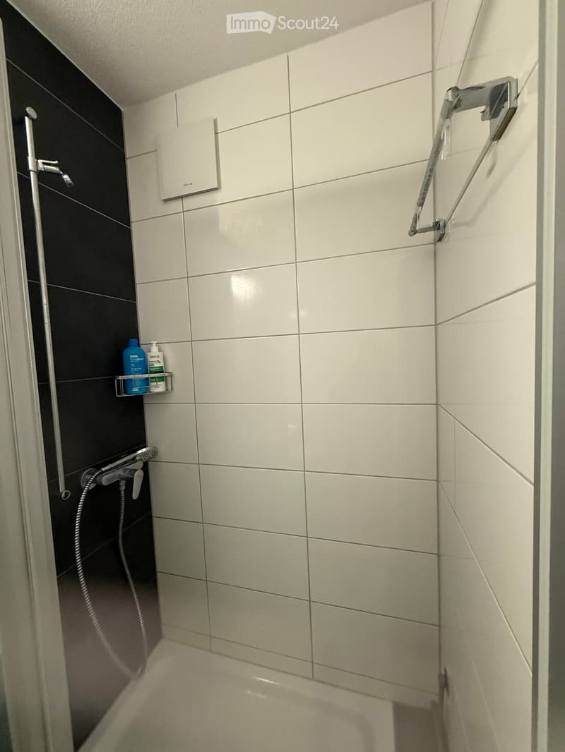 Modern shower area with white tiles, black accent wall, handheld shower head, and a small shelf holding toiletries.