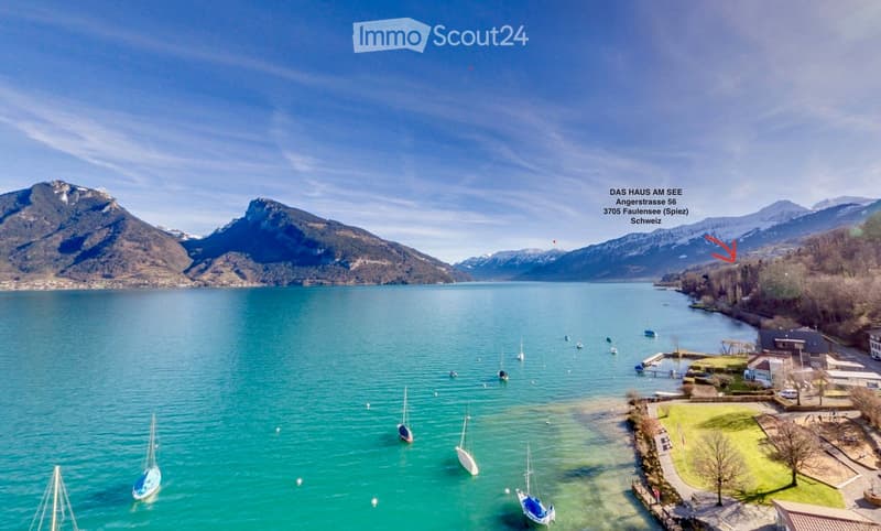 Aerial view of a serene lake surrounded by mountains, with several sailboats anchored in the water and a small dock area on the right side.