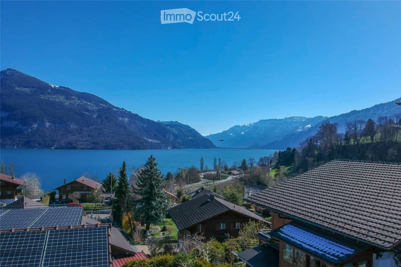 A scenic view of a lake and surrounding mountains, with several houses and solar panels visible in the foreground.