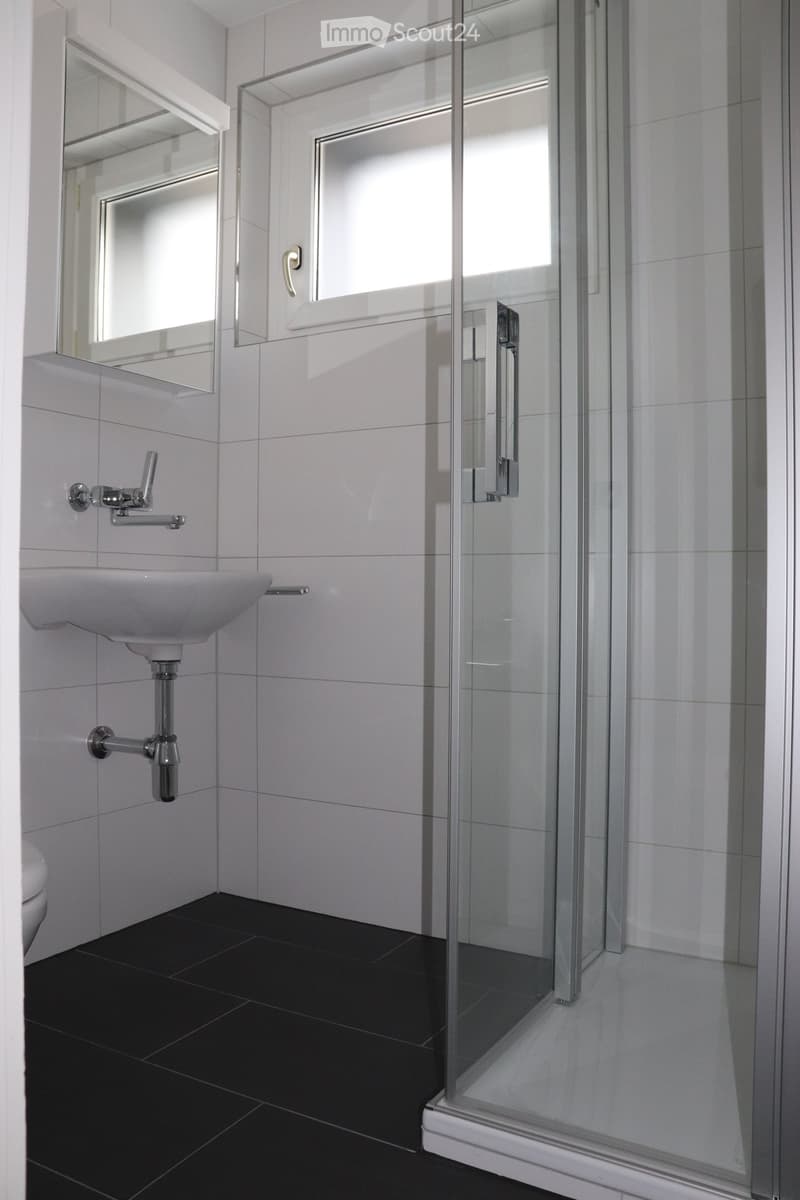 Modern bathroom with white tiles, black floor, glass shower stall, sink, and mirror.