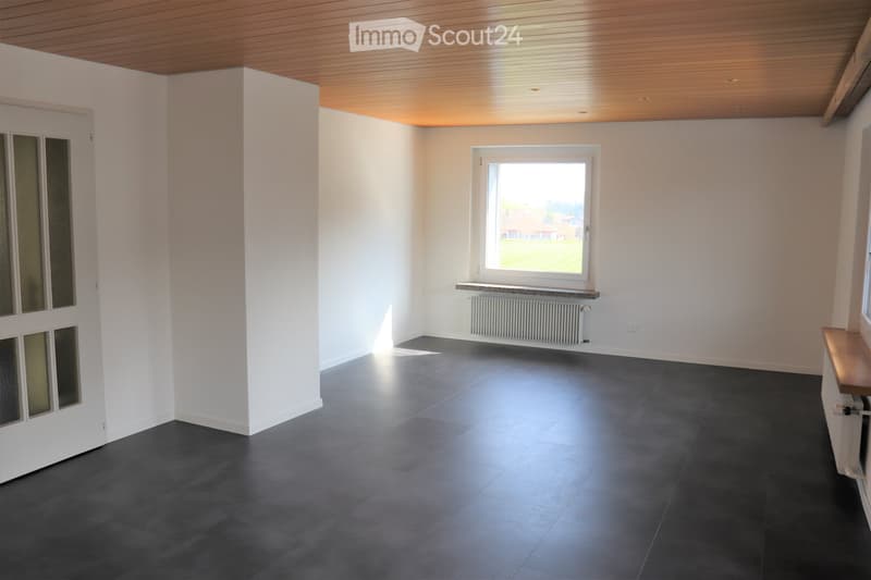 Empty room with white walls, dark grey tiled floor, wooden ceiling, and a window with a radiator below it.