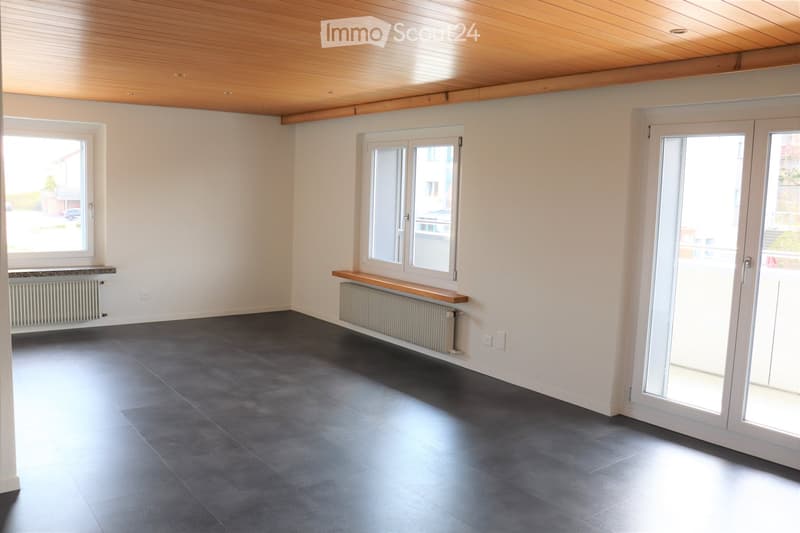 Empty room with white walls, dark grey tiled floor, large windows, sliding glass doors, and wooden bench along one wall.