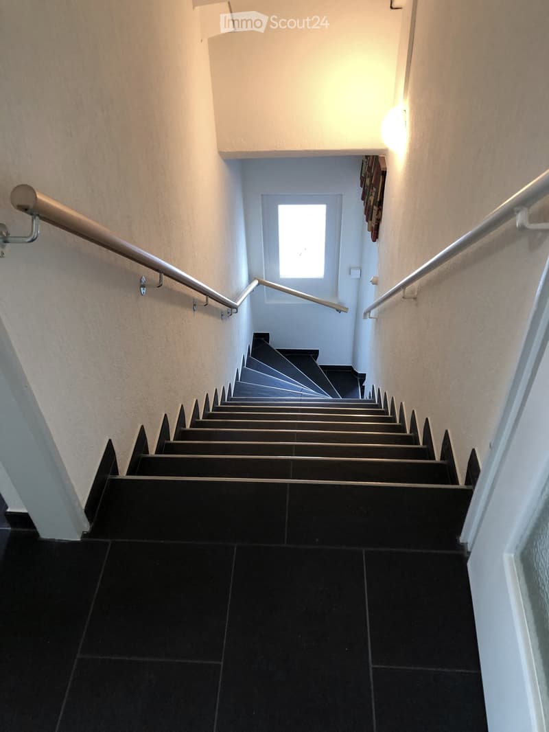 Modern staircase with black tiled steps and metal railings, illuminated by warm wall-mounted lighting, leading to a bright window at the top.