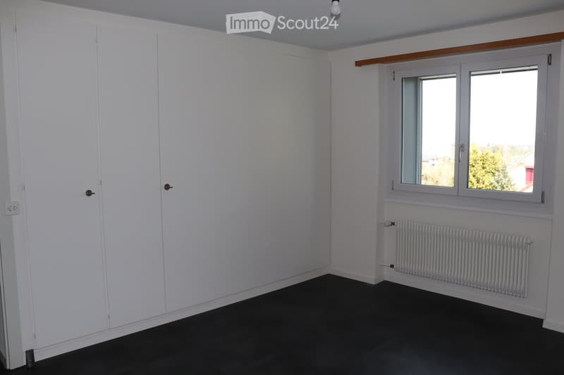 Empty room with white walls, dark flooring, and a window with white frames. The room features a radiator below the window and a door on the left side.