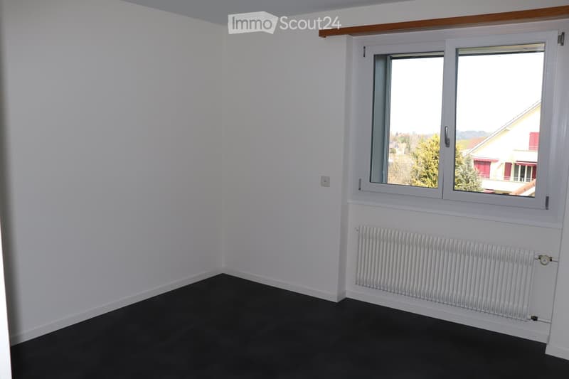 Empty room with white walls, dark flooring, and two windows offering a view of the outside. A radiator is visible under one window.