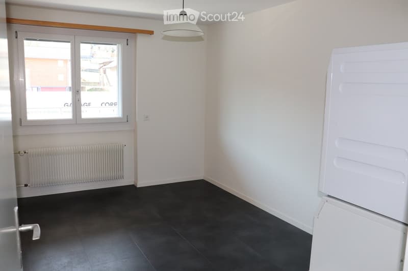 Empty room with white walls, dark tiled floor, a window with white frame, a radiator, and a white refrigerator partially visible on the right side.