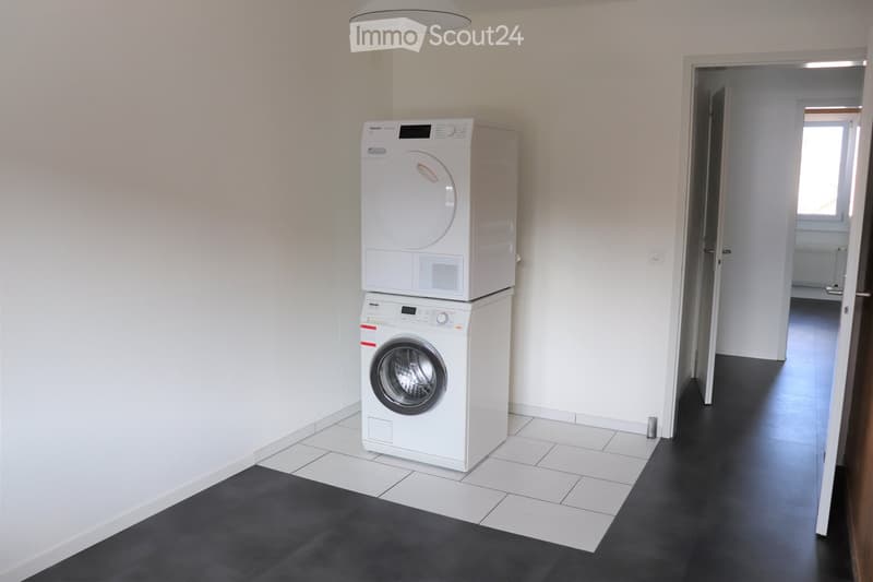 A small, well-lit room with white tiled walls and a dark wooden floor. A white washing machine and dryer are stacked against the wall, with a doorway leading to another room visible in the background.