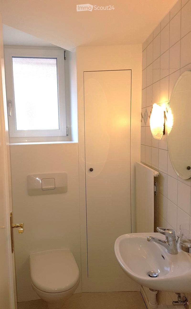 Modern bathroom with white tiled walls, a toilet, a sink with a chrome faucet, and a mirror above the sink. The room features a frosted window and a light fixture on the wall.
