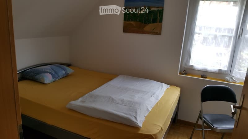 single bed with pillows and blanket, folding chair near window, white walls