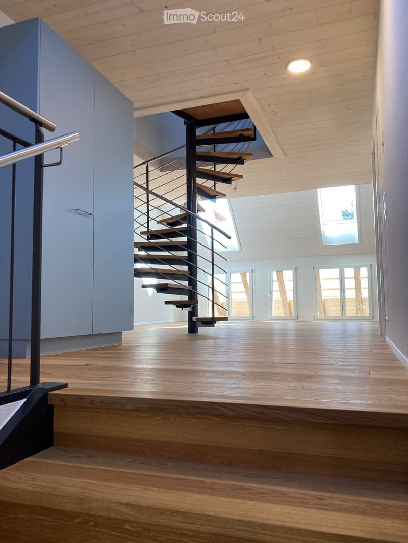 Modern, open-concept hallway with wooden flooring, exposed wooden ceiling beams, and a sleek staircase with metal railings leading to an upper level.