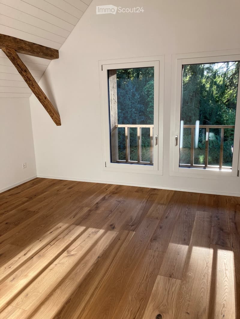 Empty room with wooden flooring, white walls, and large windows offering a view of greenery outside. The room features exposed wooden beams on the ceiling and a simple, clean design.