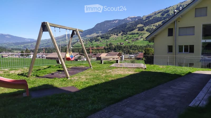 A grassy outdoor area with a swing set, slide, and bench, surrounded by a metal fence and overlooking a scenic mountain landscape.