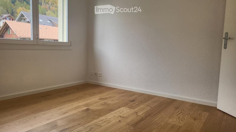 Empty room with light wooden flooring, white walls, and a large window showing a view of neighboring houses.