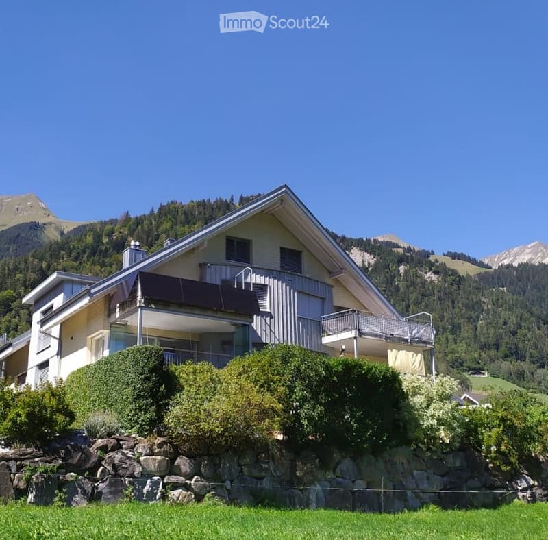 Two-story house with a balcony, surrounded by greenery and mountains in the background.