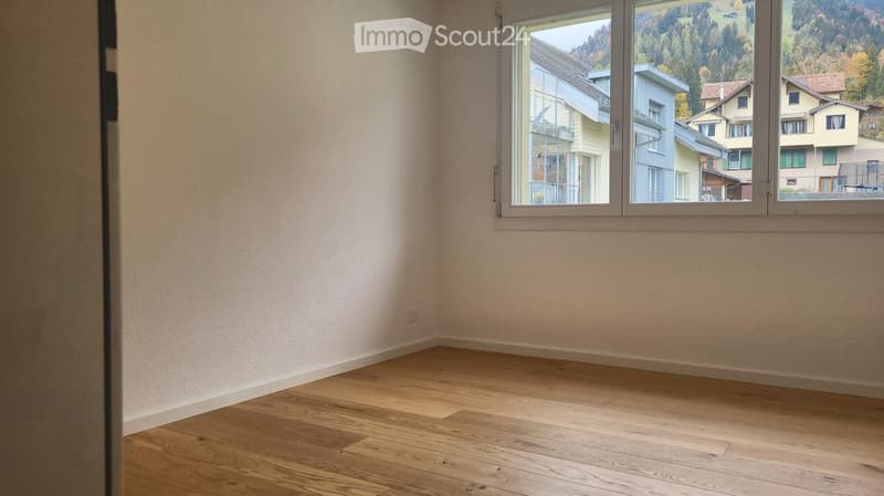 Empty room with light wooden flooring and white walls, featuring a large window with a scenic view of neighboring houses and greenery.
