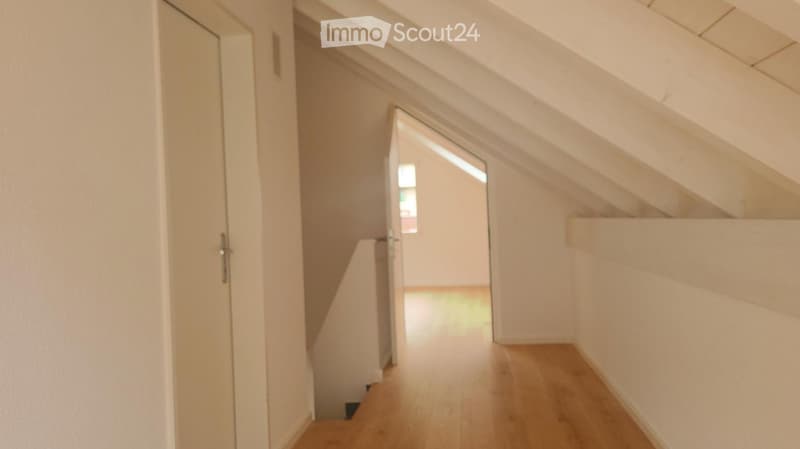 A narrow hallway with wooden flooring, white walls, and a door leading to another room. The ceiling has exposed wooden beams.