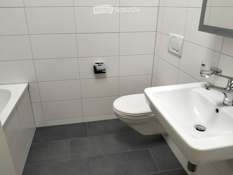 Modern bathroom with white tiles, a toilet, a sink with a chrome faucet, a mirror, and a glass cup on the sink.
